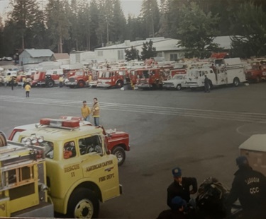 Fire engines lined up in a parking lot with firefighters gathered nearby.