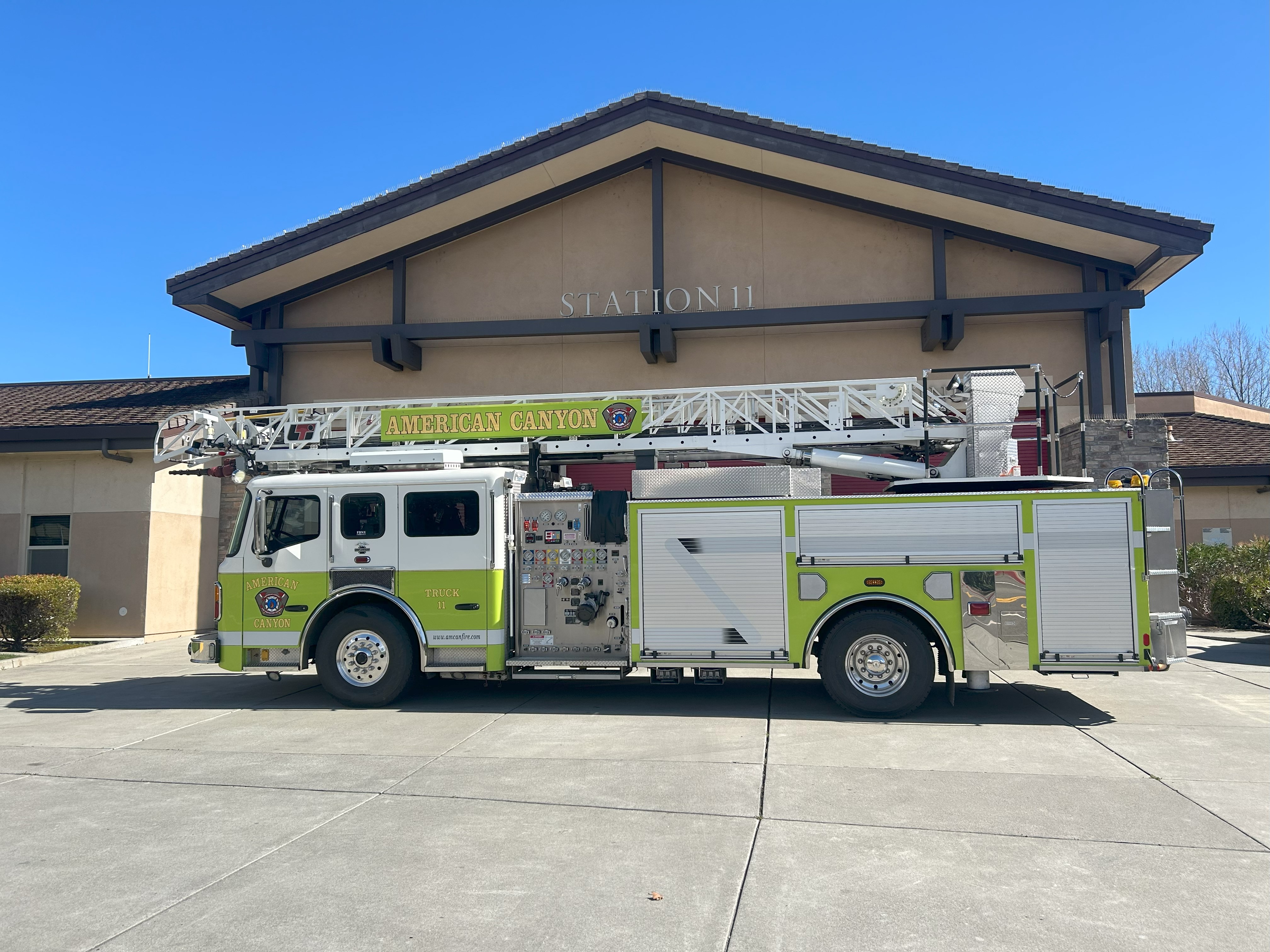 Green-and-white fire engine parked on a concrete driveway in front of a fire station building on a clear day.