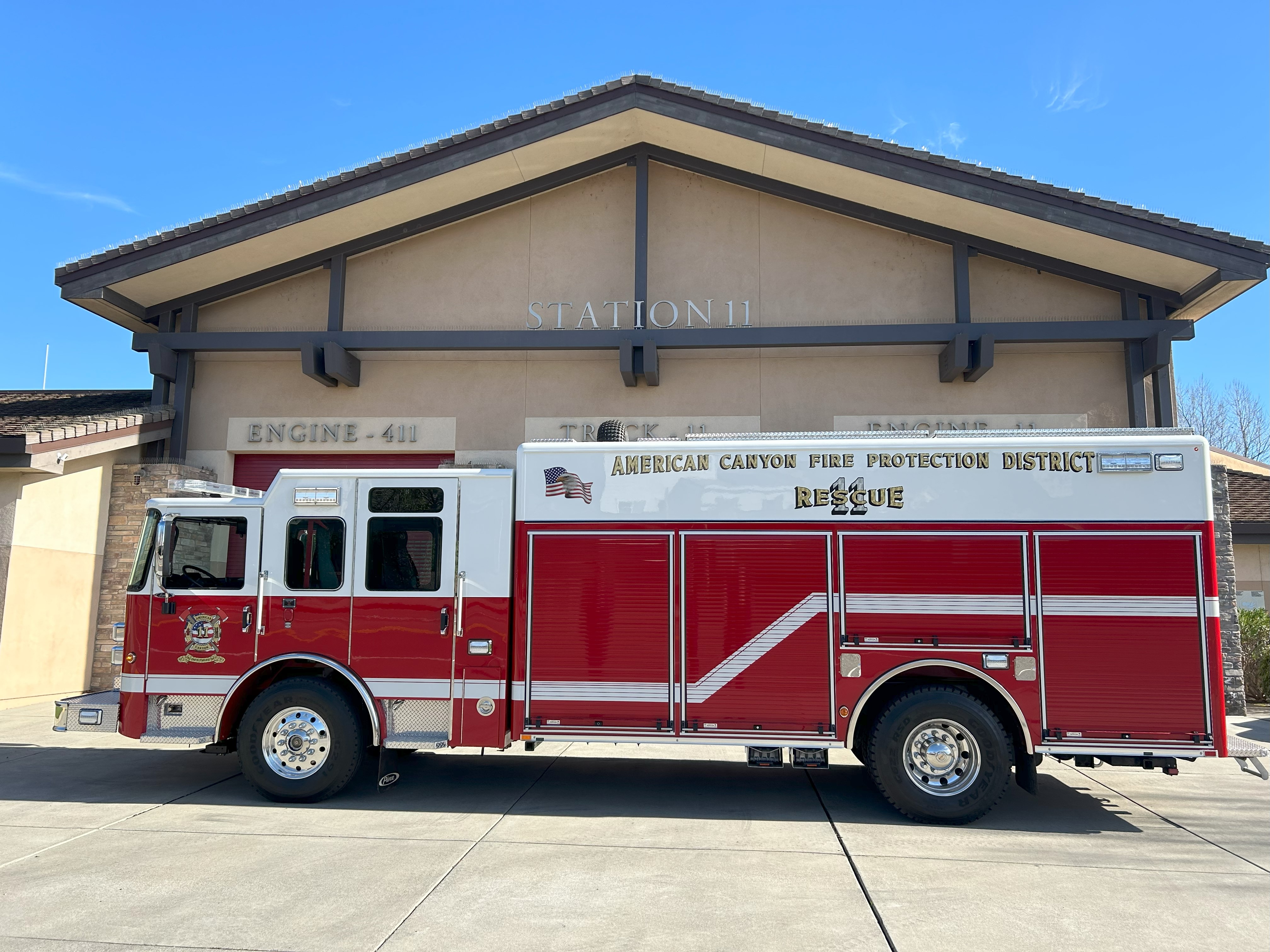 Red fire engine labeled “American Canyon Fire Protection District” parked on a concrete driveway in front of a fire station building under a clear blue sky.
